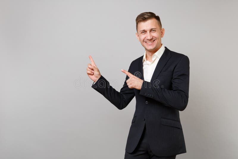 经典黑衣服的,衬衣指向食指的微笑的年轻商人画象在旁边隔绝在灰色 免版税库存图片 经典黑衣服的,衬衣指向食指的微笑的年轻商人画象在旁边隔绝在灰色 免版税库存图片